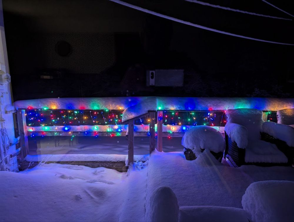 A porch railing, with multicolored Christmas lights. Some of the lights are buried under snow and are causing a diffuse glow through the snow.