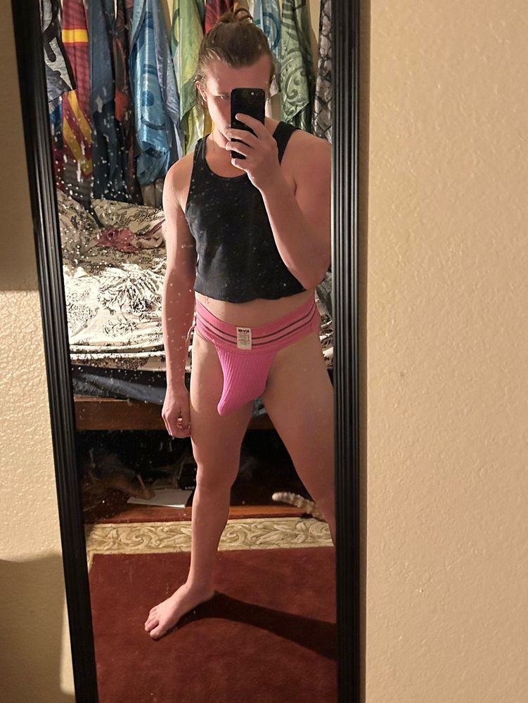 Urfaveblouse in a hot pink jock clearly very full of a very hard cock. They’re also wearing a cropped black tank top exposing their midriff and have their hair tied back, with their phone covering their face