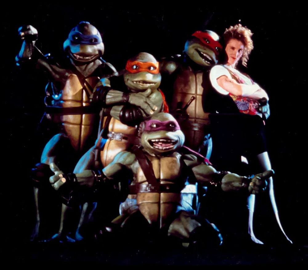 The teenage mutant ninja turtles and April from the first live action movie in the 90’s posing together smiling
