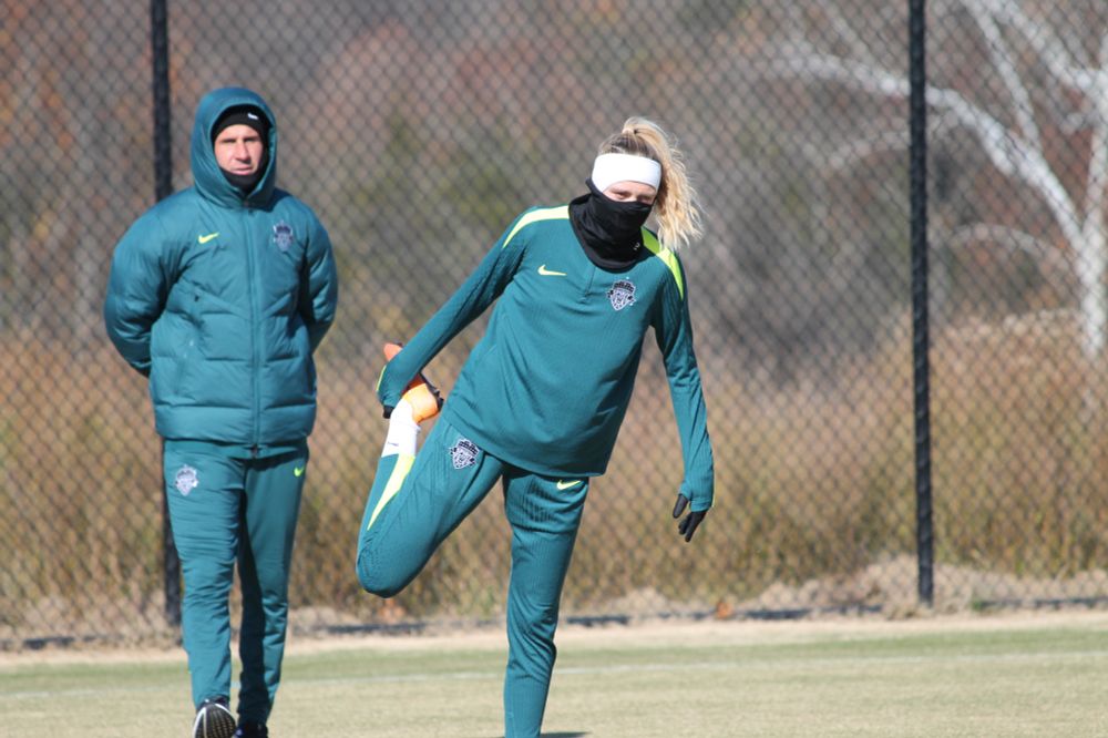 Morgan at center, stretching her right leg behind her while wearing a black face covering and a white ear-warming headband