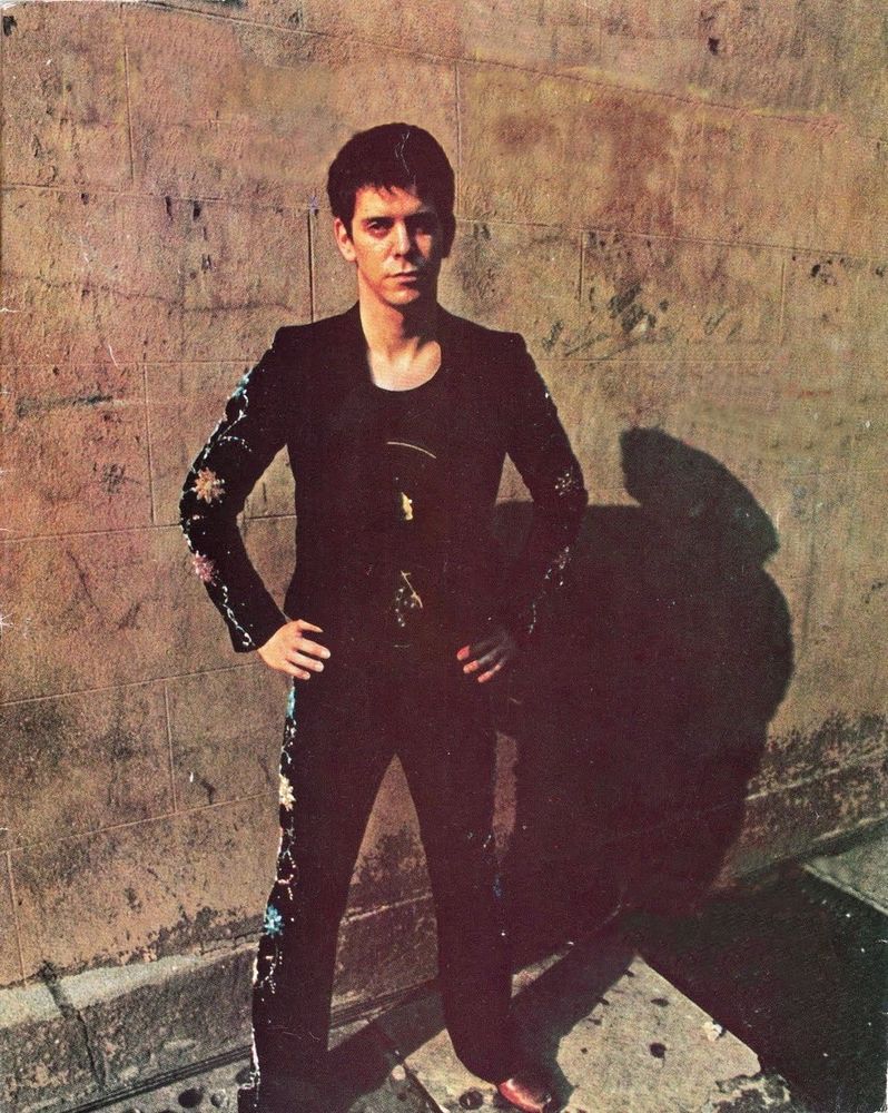 Lou Reed standing against a brick wall in tight black suit embroidered with flowers