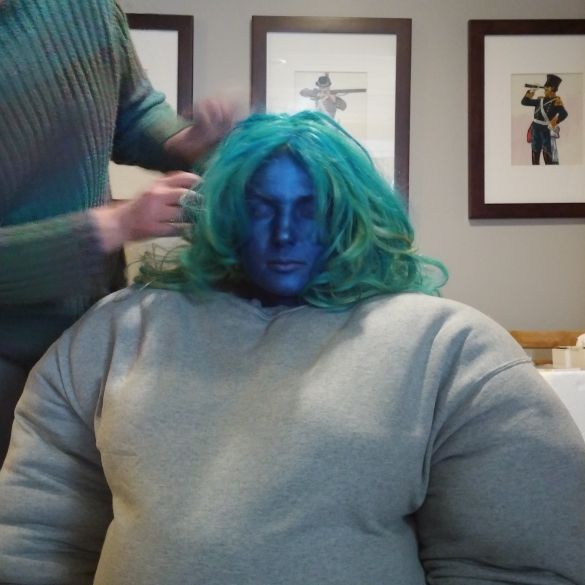 Padded girl with blue face paint and teal hair being helped by a costumer.