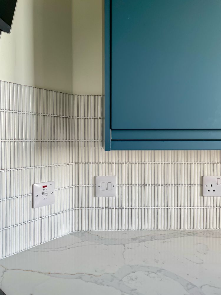 The 45 degree corner of a kitchen with part of a dark blue cabinet showing, white small vertically orientated tiles and a calacatta stone counter top.