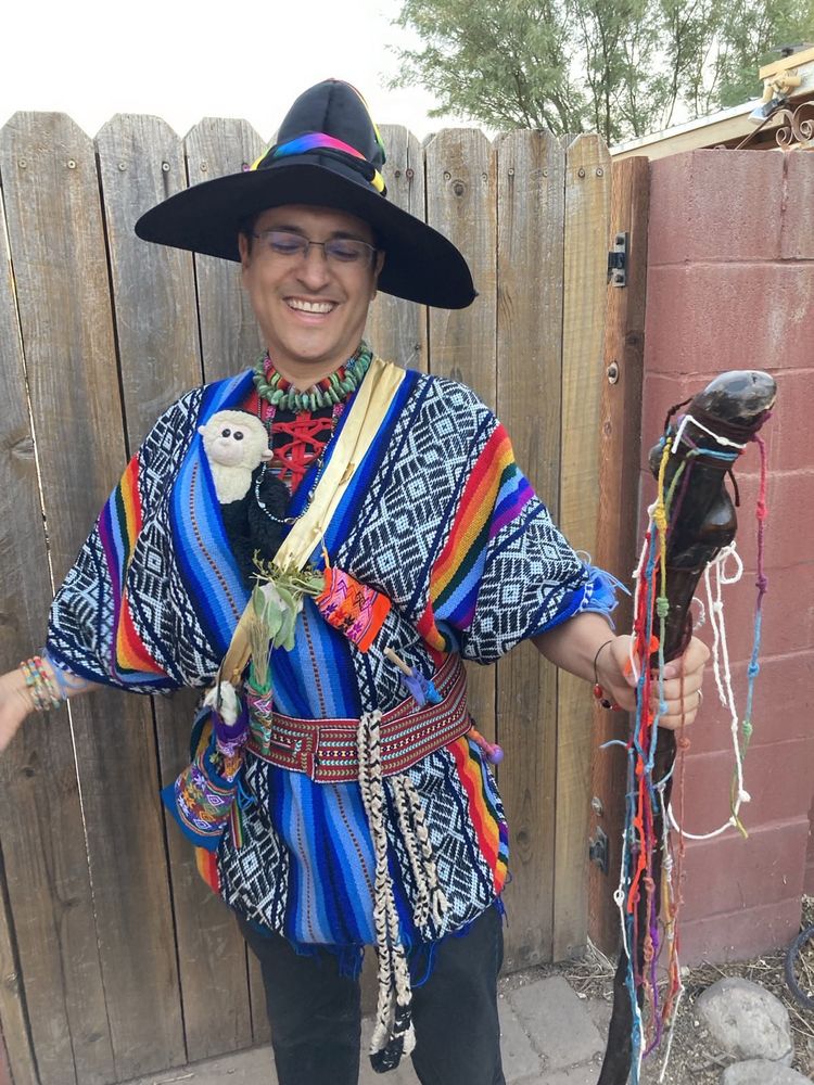 Me smiling with eyes closed and arms spread, wearing a black wizard's hat decorated with rainbow ribbon and otherwise in Quechua garb - blue pocho with red belt. A black-and-white wool sling and a wooden wand with rainbow colors in the belt. Gold sash across my front holding little bags in bright patterns holding pieces of plants, feathers, and wool.  A dark wood staff covered with knotted cords in many colors. Face and chest of a white-faced capuchin monkey are poking out from the poncho at the level of my chest.