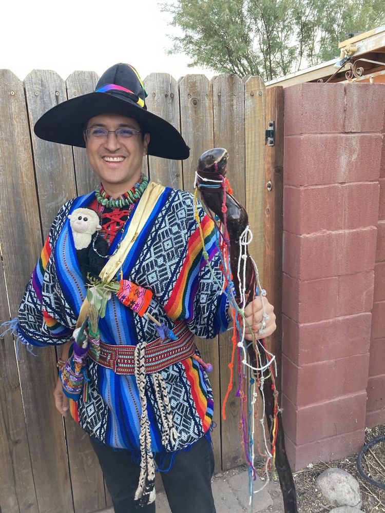 Me smiling wearing a black wizard's hat decorated with rainbow ribbon and otherwise in Quechua garb - blue pocho with red belt. A black-and-white wool sling and a wooden wand with rainbow colors in the belt. Gold sash across my front holding little bags in bright patterns holding pieces of plants, feathers, and wool.  A dark wood staff covered with knotted cords in many colors. Face and chest of a white-faced capuchin monkey are poking out from the poncho at the level of my chest.