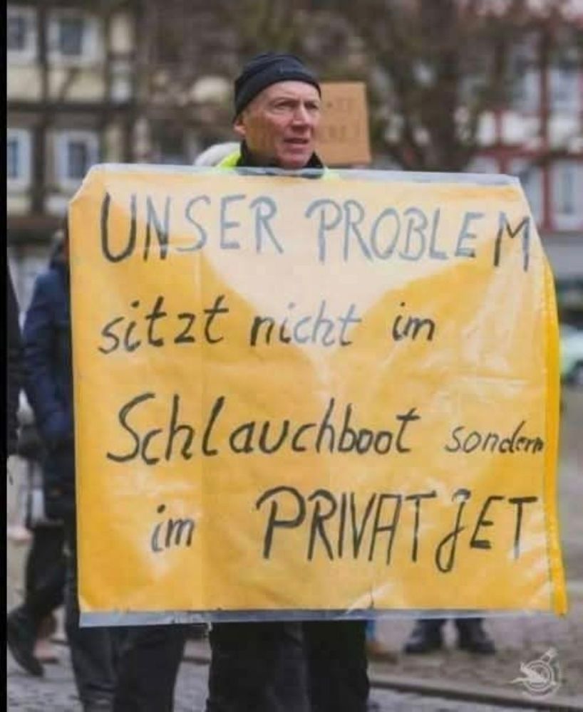 Ein Mann hält ein Schild hoch, gelb mit schwarzer Schrift, auf dem steht: Unser Problem sitzt nicht im Schlauchboot sondern im Privatjet