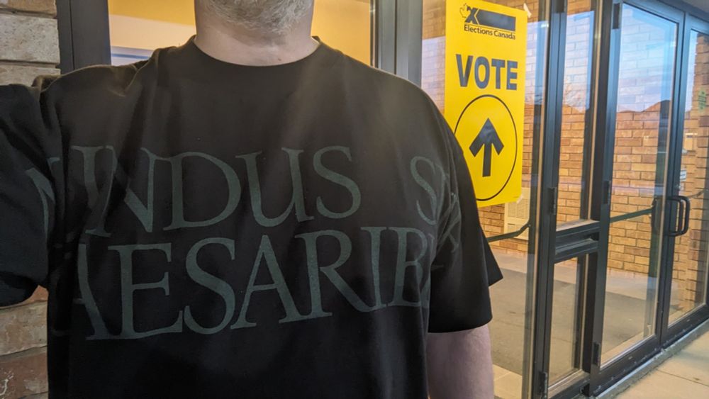 Torso of a man (me!) in a black shirt that reads Mundus Sine Caesaribus in dark text, standing in front of glass doors with a yellow sign that reads VOTE.
