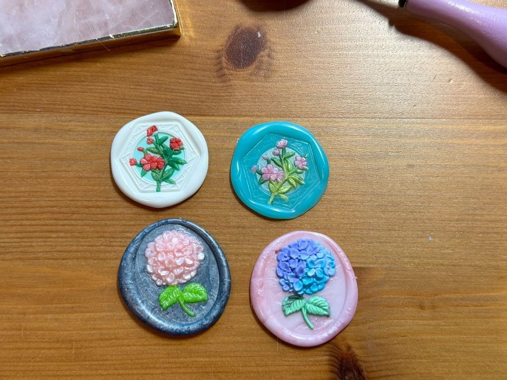 4 decorative wax seals. 2 of a stem of delicate flowers with hexagonal frame shape around them. One is red and green with a soft blue background and white frame. One is pink and green with a transluscent aqua frame. The other 2 are of a hydrangea blossom. One is shimmery pink with bright green stem on a shimmery dark grey background. One is varigated purple and blue with a mottled green ste on a pink background.