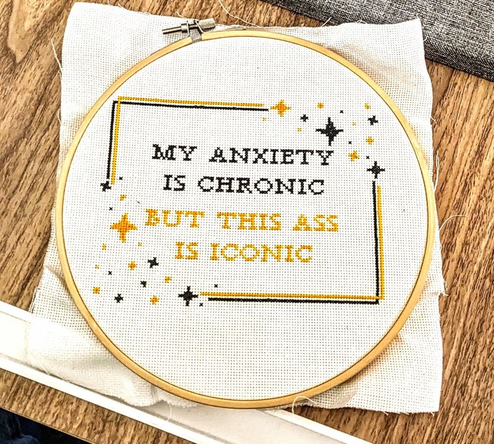 Cross Stitch piece- white Aida in hoop with black and gold thread. The pattern is a quote that says "My anxiety is chronic but this ass is iconic", with some border and sparkles as flare. 