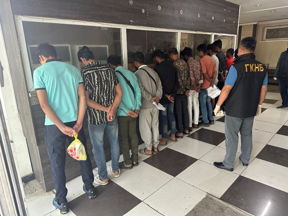 South Asian men lined up against a wall with their backs facing a Kyrgyz security services officer 