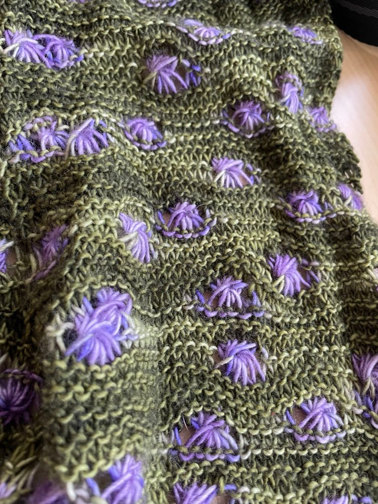 Tonal green garter stitch with bright purple aster stitches, looking like flowers on a field of green. 