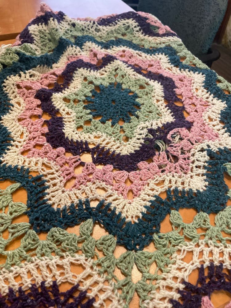 Crocheted star blanket in concentric stripes of blue, green, white dark purple, pink, and then white again before it repeats. 