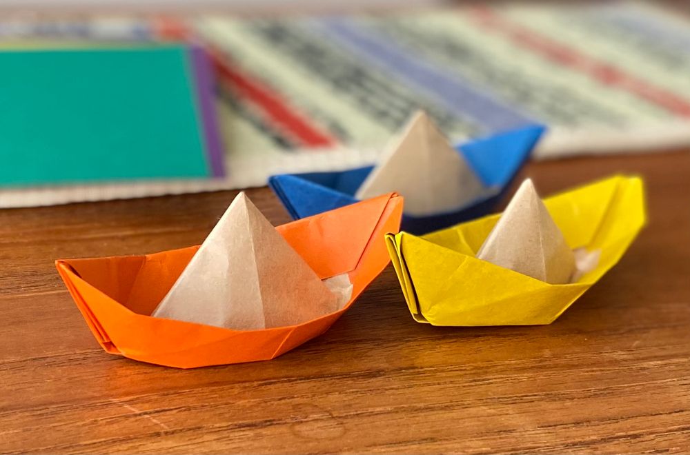 明日のスタンディングの折り紙ワークショップで折る予定の荷物船を練習中。オレンジ、黄色、青の船がテーブルに並んでる。