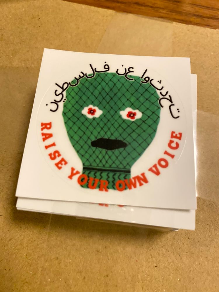 moeさん作の丸いオリジナルステッカー。グリーンのクフィーヤ柄の人の顔に、上半分にアラビア語で『パレスチナ』下に『声をあげよう』と英語で書いてある。