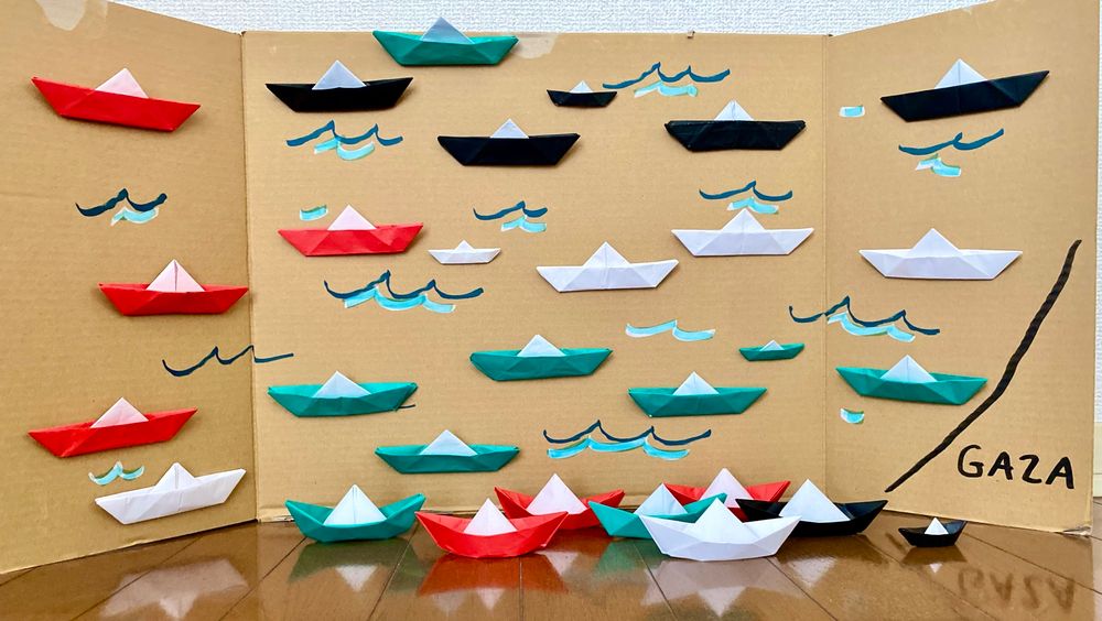 高円寺スタンディングのワークショップで作った折り紙のフロティラ船団！みんなの願いが込められている。ダンボールに波の模様と右下にガザ、そこに折り紙で作った船が貼られている。床にも何隻か置いてありガザに向かう様子を表現している。