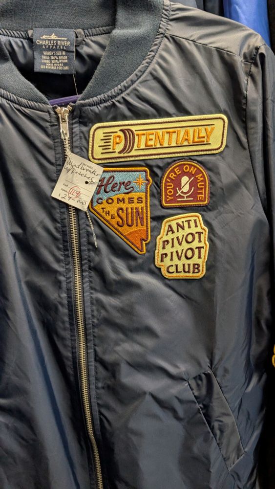The front of a navy blue jacket with patches that say: "Potentially", "here comes the sun", "you're on mute", "anti pivot pivot club"