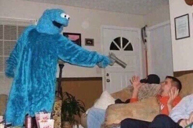 A photograph with the vibe of early 2000s digital photography, taken from a low position as if the photographer is sitting on a nearby couch. In a living room, a man sits on a couch in frame, leaning back with his hands held up and shoulders hunched in surprise, face captured in open-mouthed mid scream. Standing above him a yard away is a person in a cookie monster costume, closing much of the distance between them with a handgun held at the blue fuzzy end an outstretched arm. Cookie monster's expressionless gaping visage points downward towards the man at the couch, following an invisible line down and through the pistol, meeting the couch man's gaze. His eyes are no more than painted ping pong balls, but they embody the intrinsic chaos on display