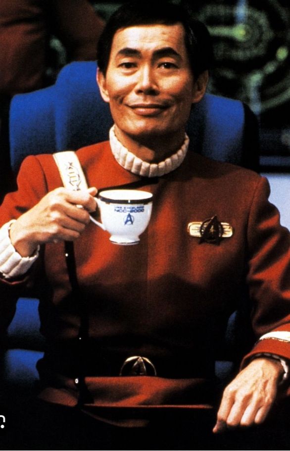 Captain Hikaru Sulu holds up a white tea cup with the words "USS Excelsior NCC-2000" written in blue above the Starfleet Delta logo. He is smiling. 