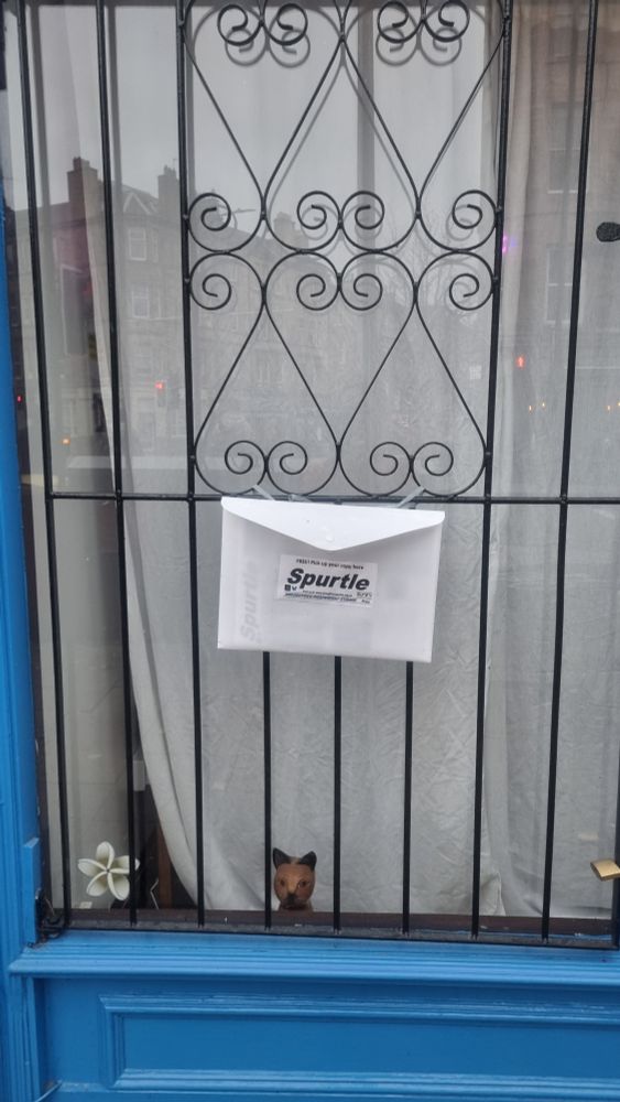 shop window with wrought iron grill. A4 poly packet is attached to the grill containing six copies of a very auspicious publication, the Broughton Spurtle.