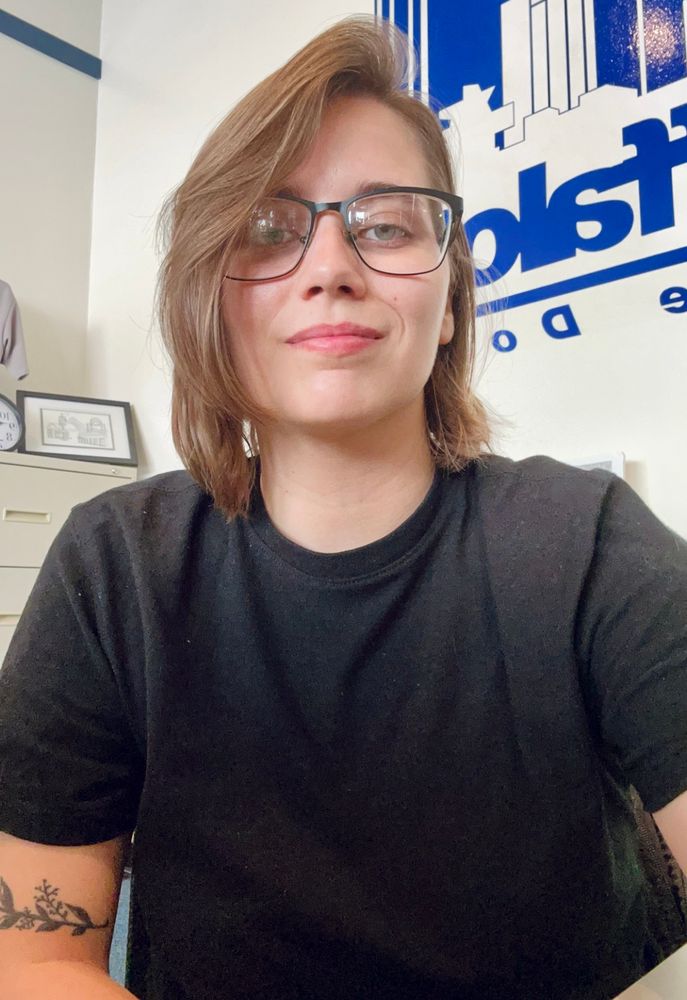 Girl with glasses on, short brown hair, black shirt, a little vine tattoo on arm 