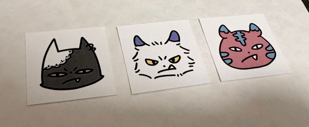 三枚の動物のイラストが描かれた正方形の紙が並んでいる写真
左から右耳に白いレース・左耳に2連ピアスをした黒猫、耳が紫・目が黄色の毛の長い白猫、ピンク地に水色のトラ模様の虎
三匹とも鋭い目つきで正面を見て牙を出している