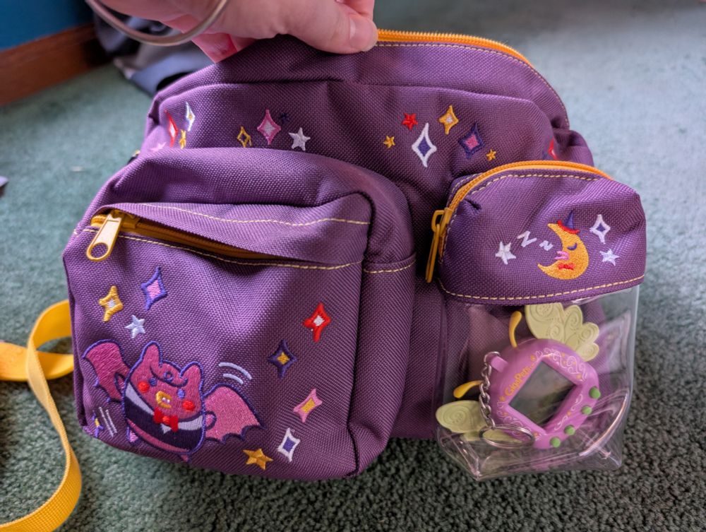 closeup of the bag, it's purple with yellow straps and has a cute sparkle design on it including a tama that looks like a bat (sorry I don't know his name). it also has a clear pocket on the front to display a plush or virtual pet device. 