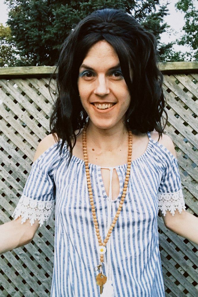 White trans woman standing on a deck smiling. She's wearing a blue and white striped mini-dress and a dark brown long wavy wig with a braid in the front