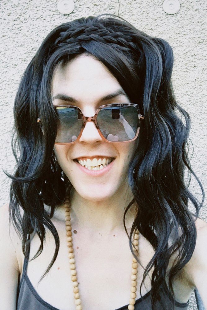 White trans woman wearing a brown camisole, sunglasses, wood bead necklace, and a long wavy dark brown wig with a braid in the front. She has a smile on her face.