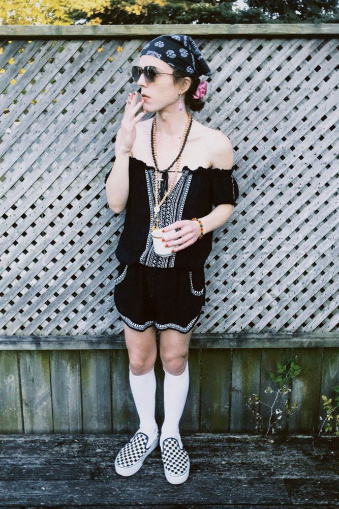 White trans woman with long brown curly hair tied back in a bun. She's standing on a deck holding a cup of coffee and smoking a spliff. She's wearing checkerboard vans platform slip-ons, white knee socks, black romper, beaded jewelry