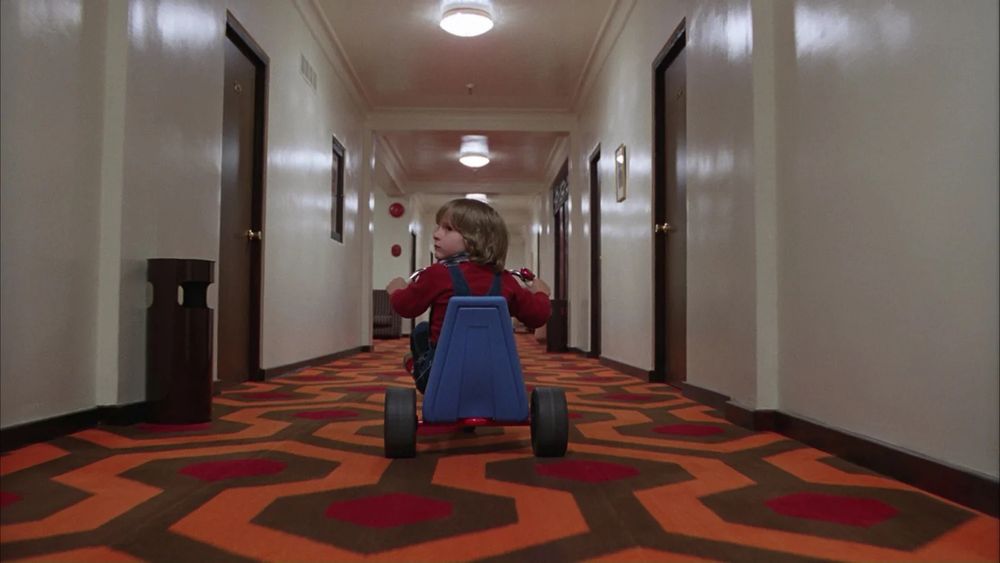 A scene from The Shining. A young boy riding a tricycle down a hotel corridor.