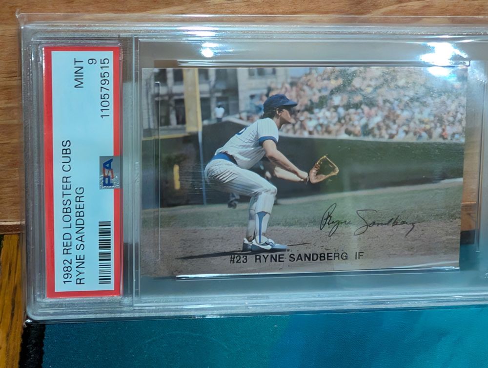 Image of a 1982 baseball card. Shows Ryne Sandberg in a fielding position at third base. He faces right in the image, looking toward home. The right field foul pole and the first base side stands (full of people) are in the background.