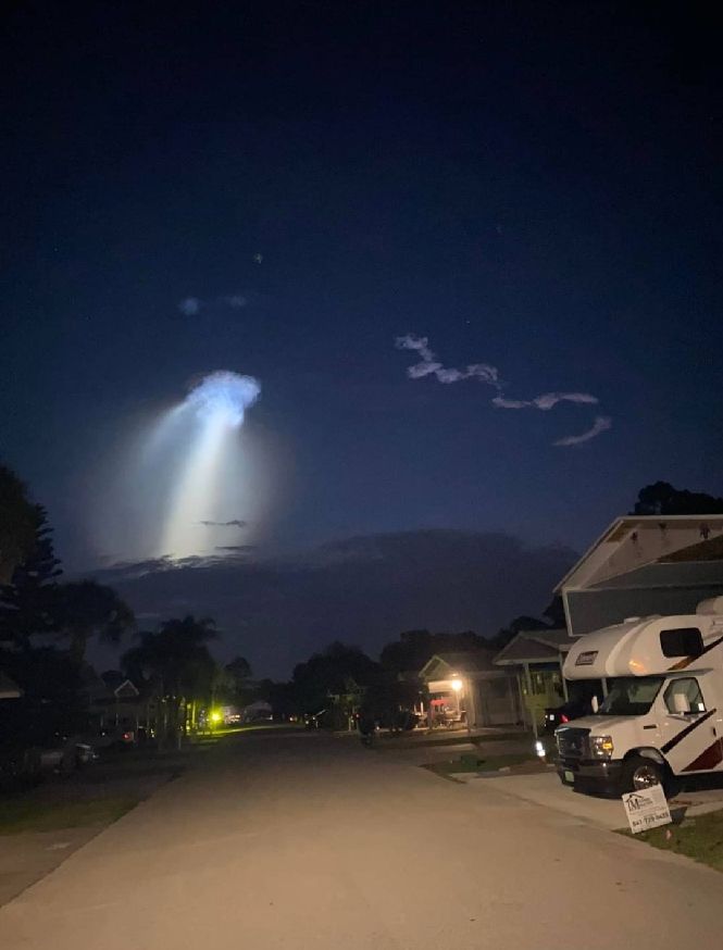 Early morning view of a street, with dark pre-dawn skies. A large plume of rocket exhaust appears in the sky, starting low with a wide white plume and then expanding at the top into a wide mushroom cap. It looks like a giant sky jellyfish.