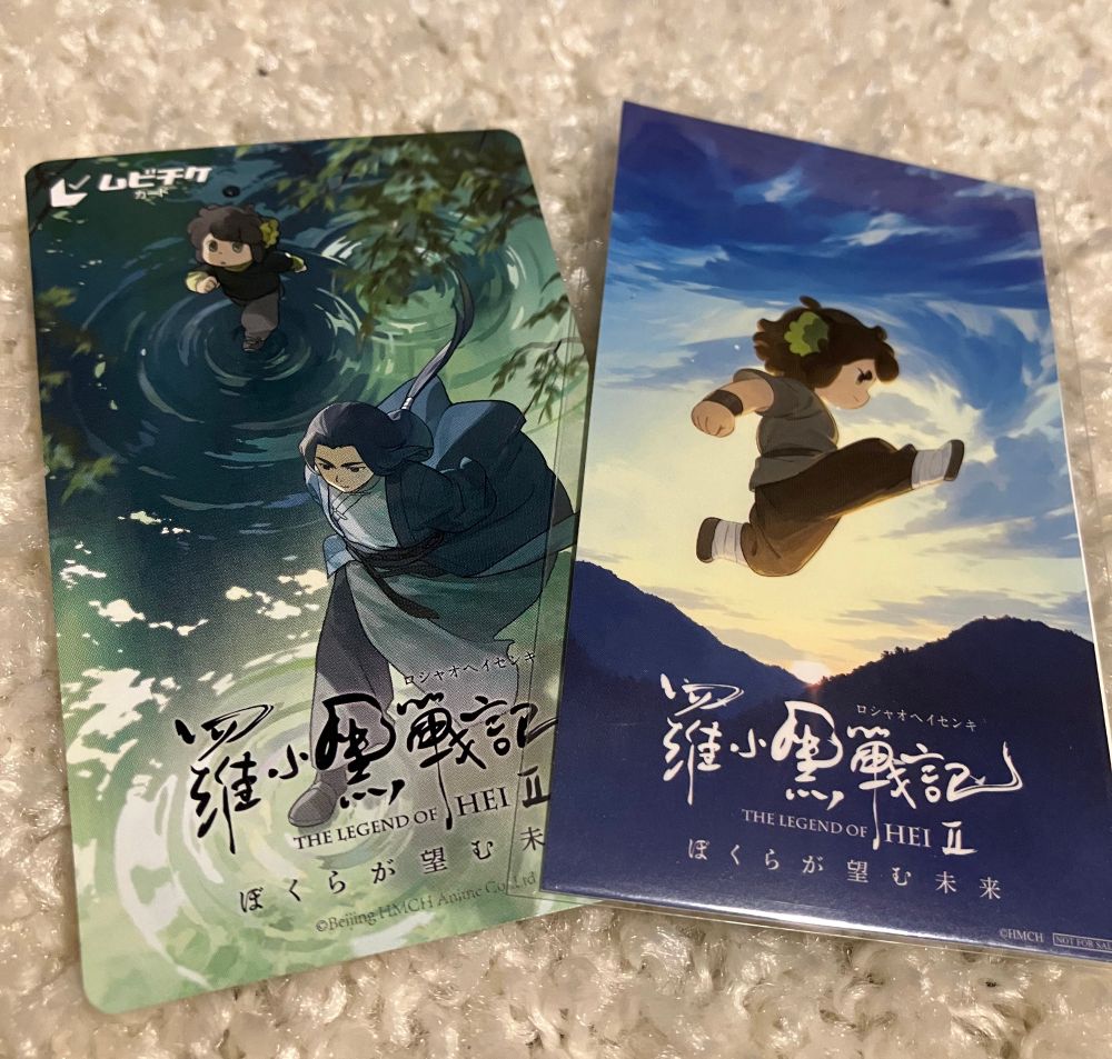 ロシャオヘイセンキツーの前売り券とステッカー。緑に光る水面を歩くムゲンとシャオヘイが描かれた前売り券。朝焼けに飛び出すシャオヘイが描かれてステッカー。