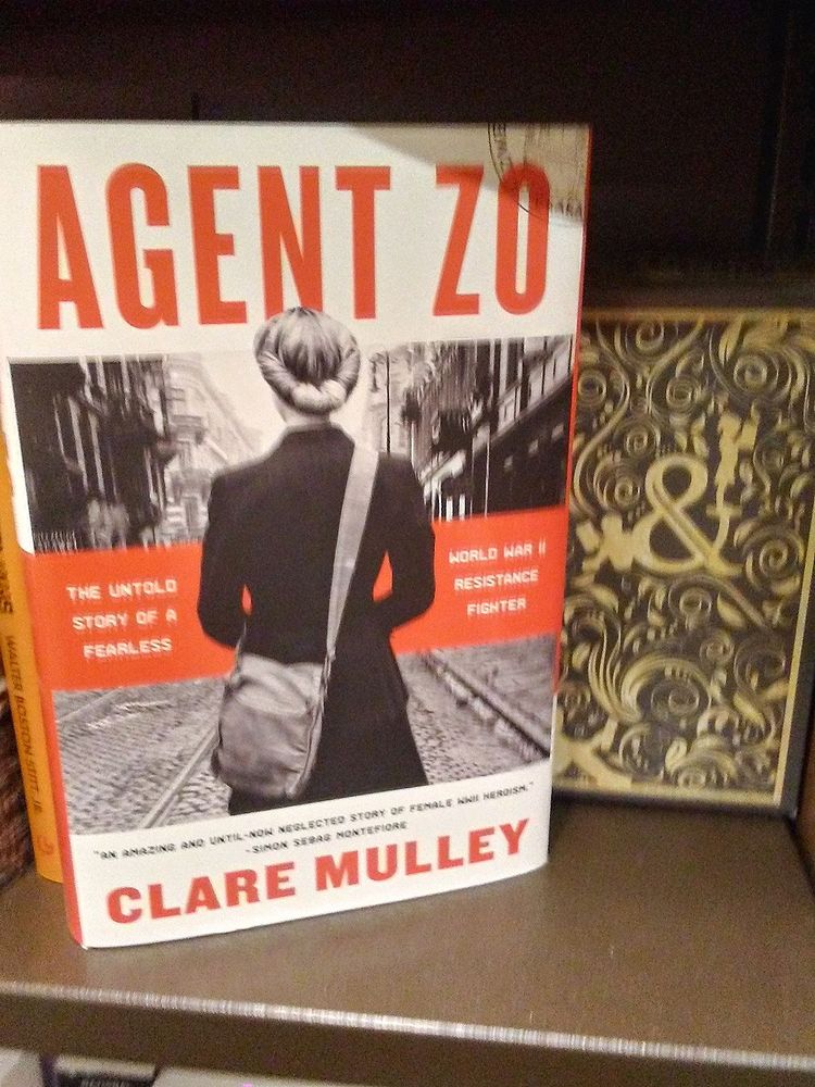 Book on a bookstore shelf cover-facing out with a decorative Barnes & Noble filler box. Cover shows a non-fiction biography of a WW2 Polish courier and resistance fighter, Agent Zo, by Clare Mulley (the title and author splashed in type across the top and bottom of the cover). The middle illustration, in black and white, shows the back of a woman in a coat and satchel slung over one shoulder walking down a European street, flanked by the words “The untold Story of A Fearless World War II Resistance Fighter.” 