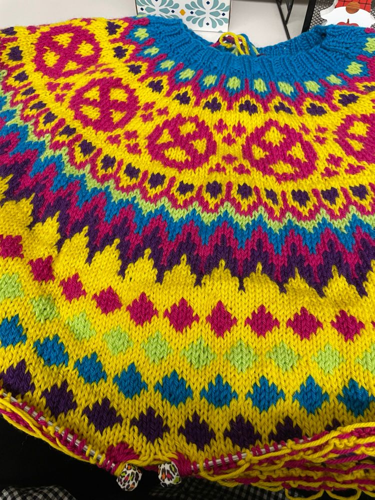 A photo of an in-progress knitted sweater in colorwork with yellow, pink, green, blue, and purple designs. The yoke of the sweater features the X-Men’s logo from the 90s animated series of a circle with an x in it.