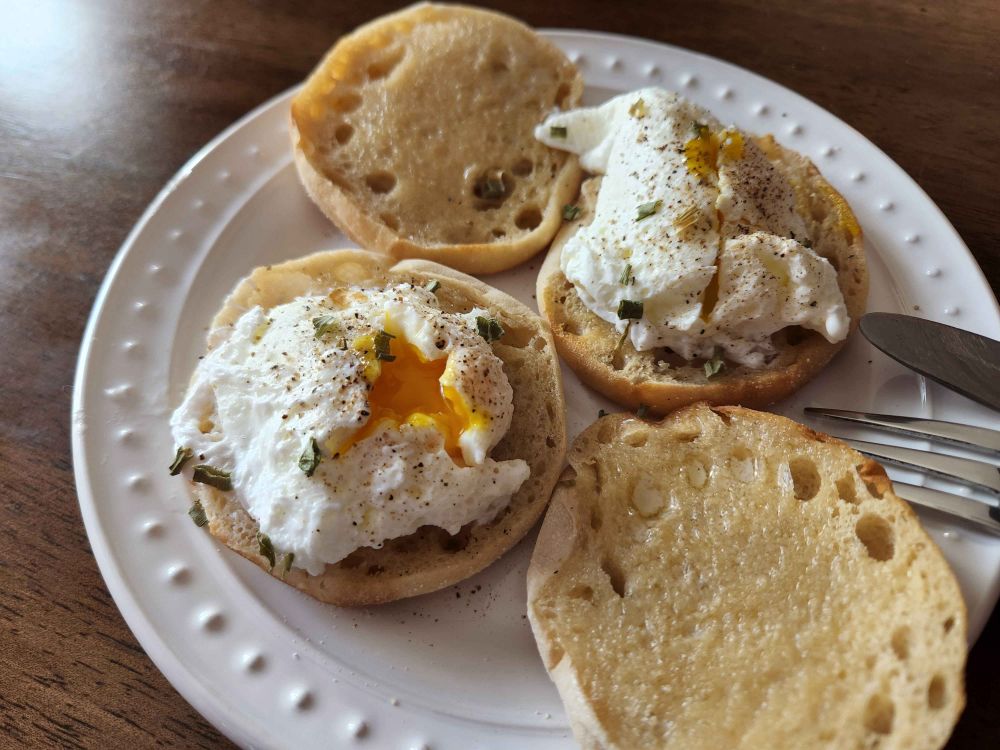 A photo of two poached eggs on buttered English muffins, on a white plate. The eggs have been garnished with dried chives and salt and pepper, and both are sliced open to reveal the runny yolks.