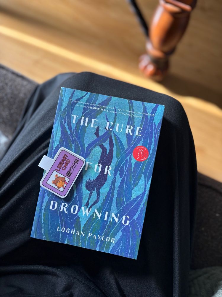 A picture of the book The Cure For Drowning on Amy’s lap with a DW Library Card bookmark on top of it