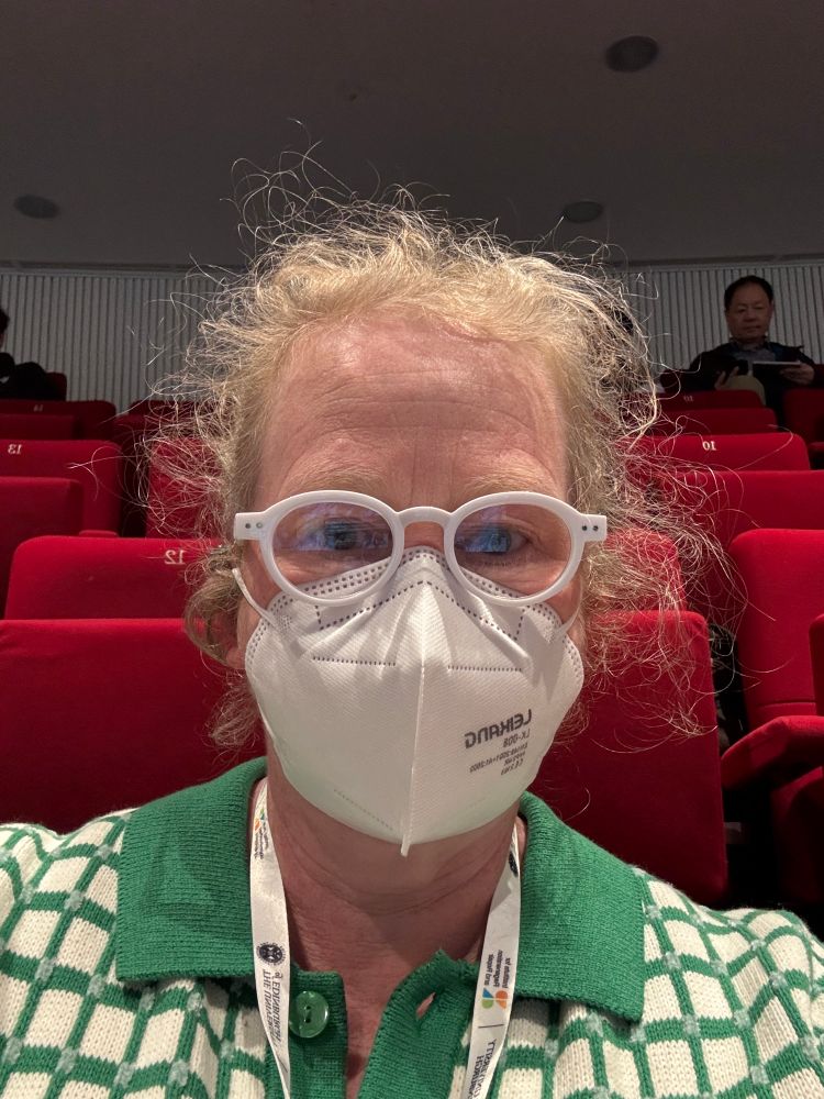 Woman in an auditorium, she is 1 of 3 wearing an FFP2, sadly that doesn’t include the woman in front of us who’s coughing…