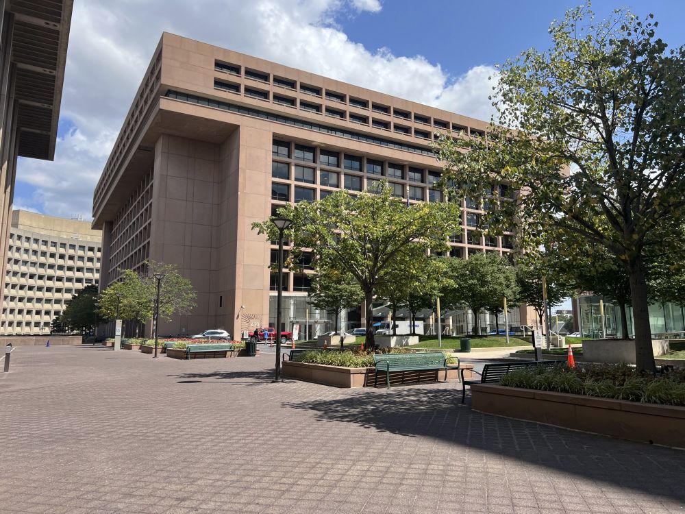 Office buildings at L'Enfant Plaza, in central DC. Image shows three concrete office buildings with glass windows and wide, empty sidewalks, with a few trees in raised tree boxes surrounded by flowers.