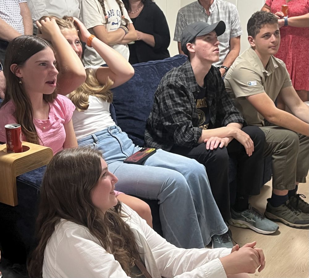 Three teenaged girls and two teen boys sit on a floor and sofa, looking at a television screen off screen. One girl has her arms raised to her head raised in an emotional response. One girl's mouth is open. One girl is smiling. The boys are smiling. Adults can be partly seen standing behind them.
