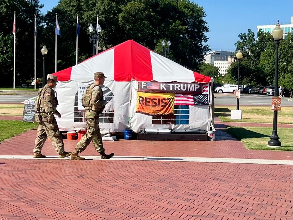 Two yound men in khaki camouflage, branch of service unclear, pass in front of a messy red and white tent with 