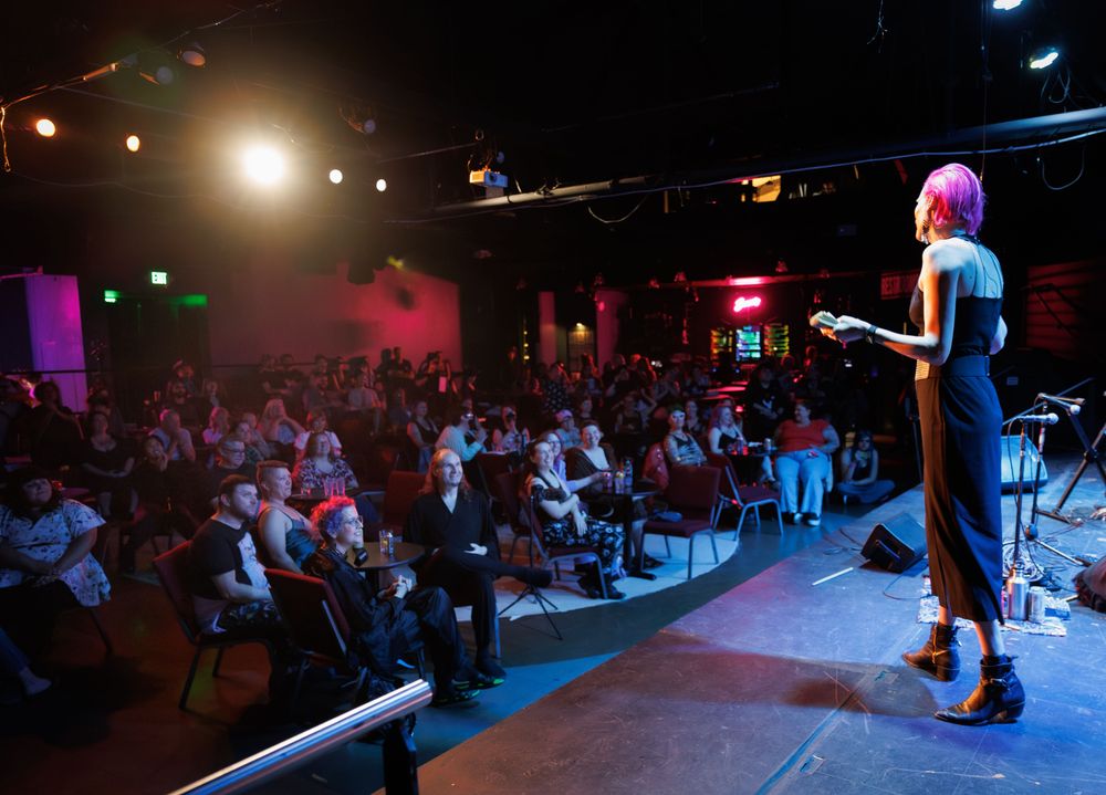 Allie, a tall pink-haired women in a long black dress, stands on stage in front of a packed theater of smiling faces. Vibrant multi-colored lights around the theater make the photo feel alive
