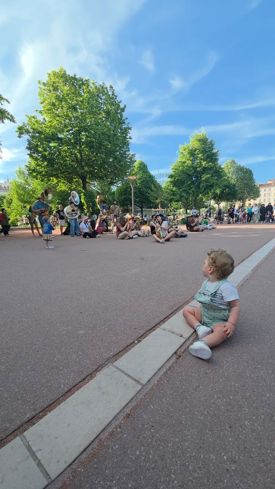 Notre fils est assis de trois quart dos avec une salopette verte d'eau il regarde une fanfare en arrière plan. Il y a également des arbres et un beau ciel bleu.