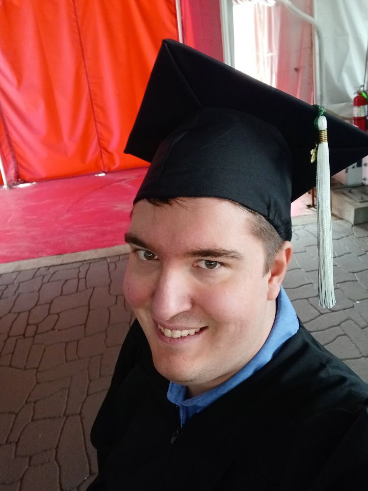 A white person with brown hair wearing a blue shirt and black cap and gown with green and white tassel standing against a red curtain and cobblestone floor. They are smiling.