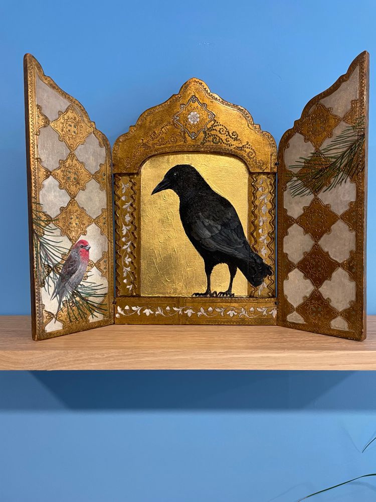 Triptych with a crow pained on gold background in the center panel, a house finch sitting on a sprig of ponderosa on the left panel toward the bottom, and another ponderosa sprig on the right panel near the top. The side panels also have a gold and silver diamond pattern covering them. 