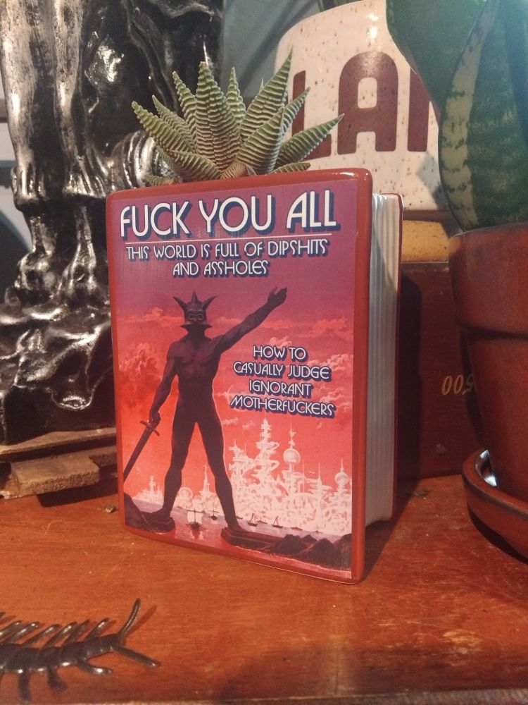 In foreground a plant holder shaped like a book has a cover with some figure brandishing a sword with an apocolyptic looling background. Book cover reads 'Fuck you all. The world is full of dipshits and assholes. How to casually judge ignorant motherfuckers'.