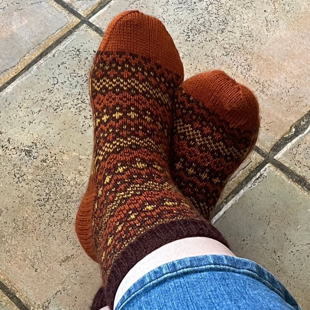 A pair of hand-knit socks on someone’s feet, resting on a tiled floor. The socks feature a warm colorwork pattern in brown garnet, orange, and yellow, with orange toes and heels, and a deep brown cuff. The intricate design has a repeating geometric motif that feels autumnal.