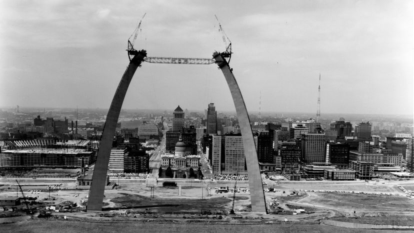 Black amd white image of an incomplete St Louis Arch. Sort of looks like 2 leaning columns with 2 special cranes mounted on top of each column. The city of St Louis sits in the background.