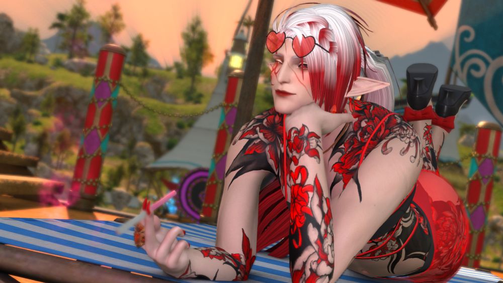 Faellont at the beach. In a black and red two-piece with a sheer red sash over his waist. He is wearing black and red heels and has heart snapped sunglasses perched onto of his head. His red floral tattoos are visible all over his body and a cigarette is between his fingers. He is winking at the camera with a soft smile.