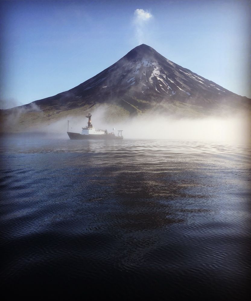 The R/V Oceanus sits offshore of Chuginadak Island while steam rises from the volcanic mountain peak.